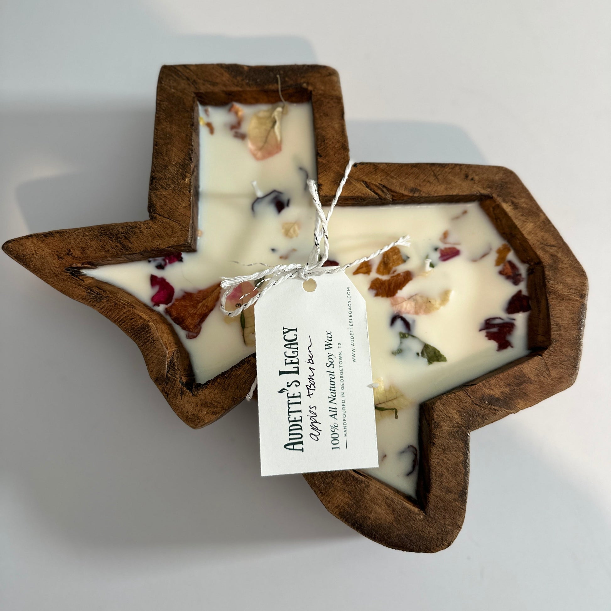 A Texas-shaped soy wax candle with a dark brown wooden frame, filled with off-white wax and dried botanicals, tied with a white paper tag that reads 'Audette's Legacy'
