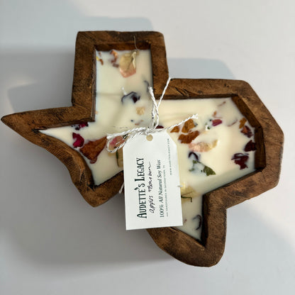 A Texas-shaped soy wax candle with a dark brown wooden frame, filled with off-white wax and dried botanicals, tied with a white paper tag that reads 'Audette's Legacy'