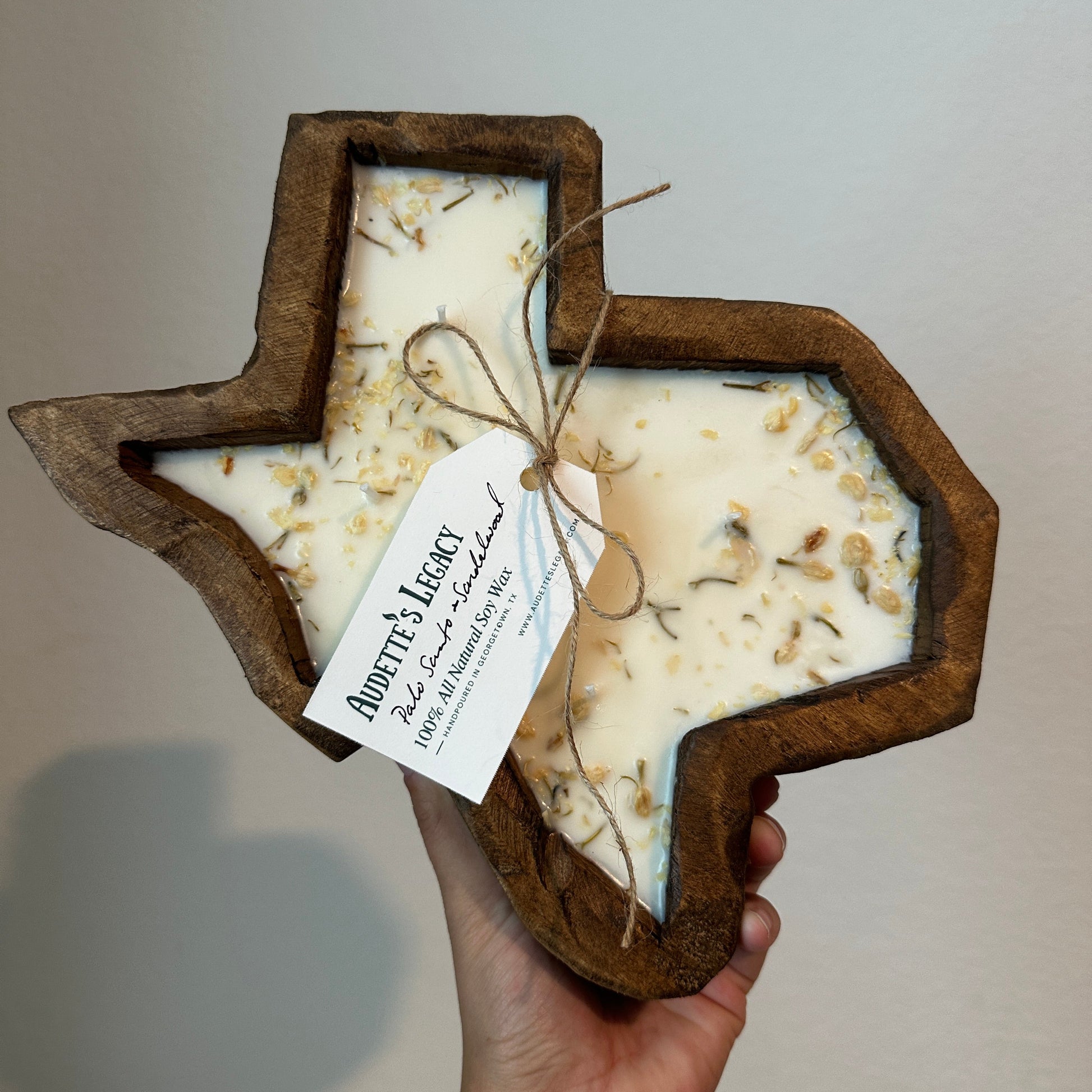 A hand holds a wooden Texas-shaped tray containing two stacked white candles with dried botanicals